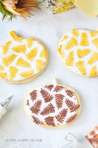 A close-up of four cookies shaped like pumpkins, each decorated with intricate fern leaf designs. Three have yellow and white icing, and one has brown and white icing. A piping bag is visible on the side, and there's a sunny, autumn-themed decoration nearby.