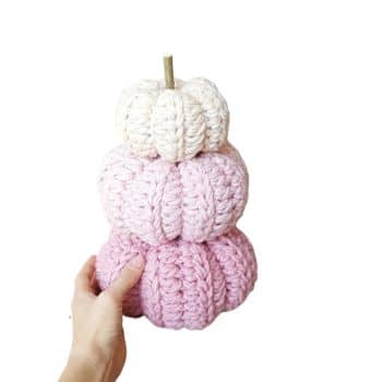 A hand holding a stack of three crocheted pumpkins in soft pastel colors. The top pumpkin is white, the middle one is light pink, and the bottom one is a slightly darker pink. Each pumpkin has a visible knit texture and a small stem on top.