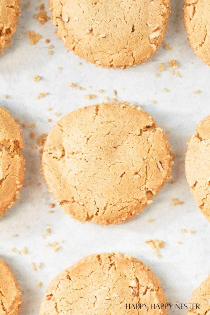 A close-up image of freshly baked cookies arranged on a light surface with scattered crumbs. The cookies have a golden-brown color and a slightly cracked texture, indicating their crispiness. The surface underneath appears to be parchment paper.