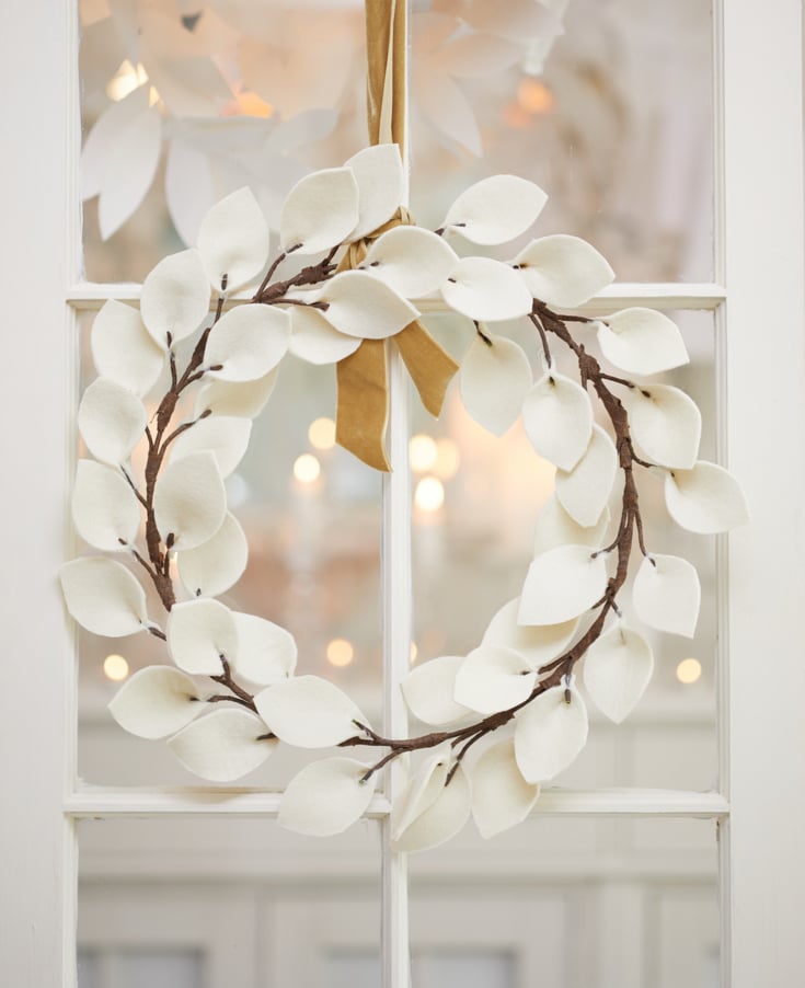 A white wreath made of soft, felt-like leaves hangs from a golden ribbon on a glass pane door. The room beyond is softly lit, with a warm ambiance perfect for fall decor ideas.