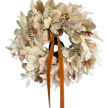 A wreath made of beige, cream, and light brown dried flowers and leaves. The wreath features small white berries and is adorned with an orange ribbon hanging in the center. The overall design has a soft, natural, and rustic appearance.