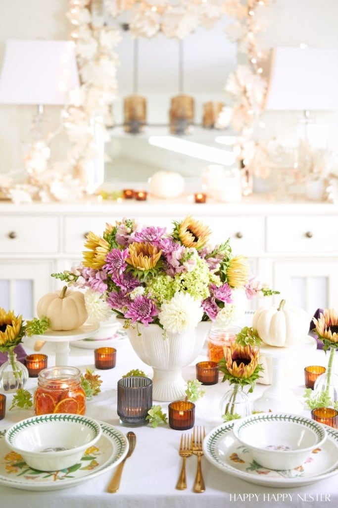 A dining table is elegantly set with fall decor ideas, featuring floral arrangements with sunflowers, green and purple flowers in a white vase centerpiece. Surrounding the centerpiece are white pumpkins, candles in glass holders, and vibrant tableware with a nature-inspired design.