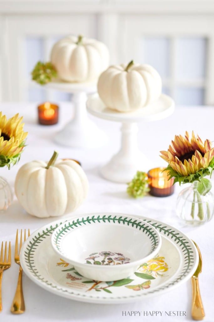 A fall-themed table setting features white pumpkins on white stands, sunflower arrangements in small glass vases, and decorative plates with green trim and floral designs. Two lit candles add a warm glow, and gold cutlery complements the autumn decor.