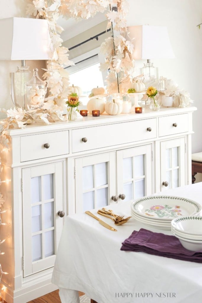 A white sideboard decorated with autumn-themed white pumpkins, ornamental leaves, and small candles. Two elegant lamps with white shades are placed on the sideboard. A dining table in the foreground is set with a white tablecloth, plates, and purple napkins this is all decor for dining room!