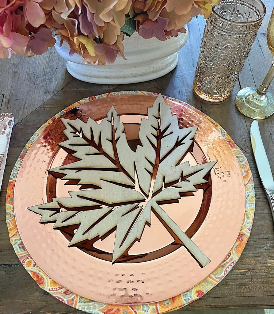 A table setting featuring a large wooden maple leaf centerpiece on a copper-colored plate with a decorative edge. The festive fall decor includes a clear, ornate glass, cutlery, and a floral arrangement in the background. The overall theme reflects an autumnal aesthetic.