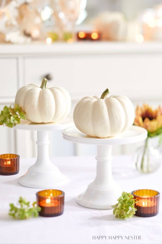 Two small white pumpkins rest on white cake stands adorned with green foliage. The setup is accented by amber-colored votive candles and a sunflower in the background, creating a cozy and elegant autumnal display on a white surface.