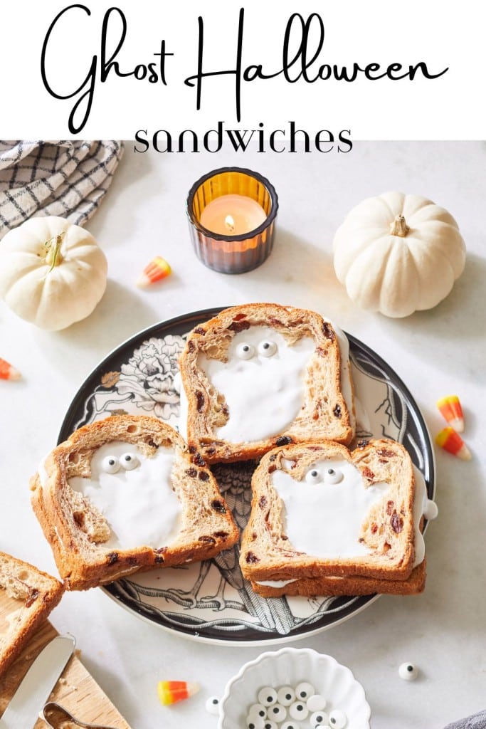 Three ghost-themed sandwiches on a plate are made from raisin bread and spread topped with candy eyes, creating a ghostly face. Surrounding the plate are decorative white pumpkins, candy corn, and a lit candle in a dark holder.