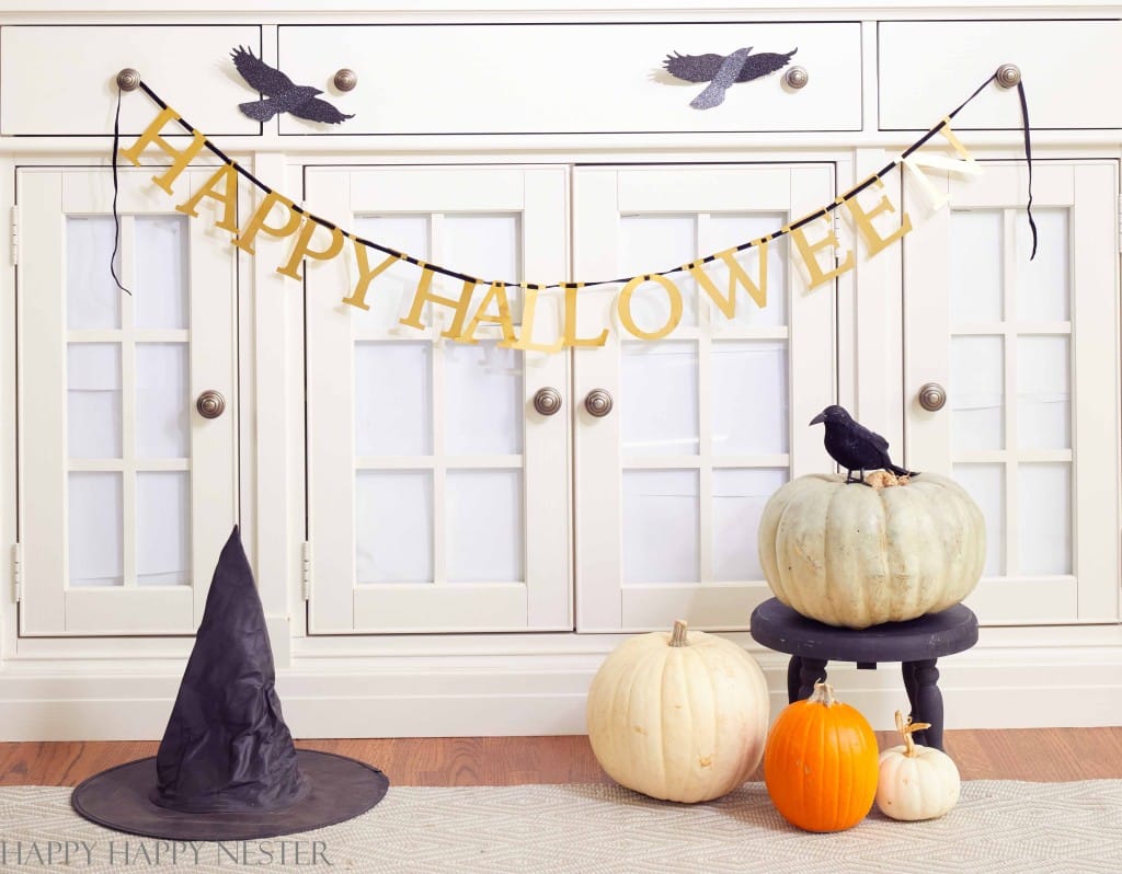 A "Happy Halloween" banner hangs on a white cabinet with crow cutouts above it. Below are pumpkins in white, orange, and a larger white pumpkin with a crow figurine on top. A black witch's hat lies on the floor next to the pumpkins and a stool, enhancing the black and gold Halloween decor theme perfectly.