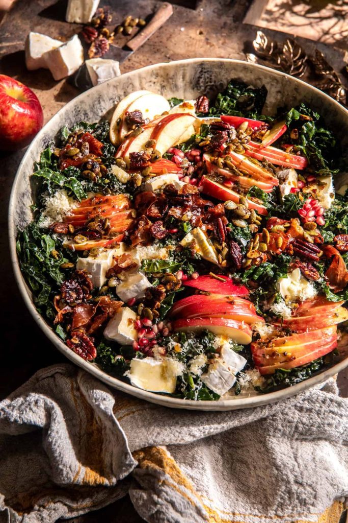 A colorful salad featuring kale, sliced apples, nuts, seeds, cheese, and pomegranate seeds is served in a rustic, textured bowl. Perfect for autumn recipes, the ingredients are arranged in a visually appealing manner with a gray striped cloth partially visible under the bowl.