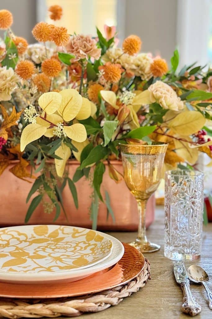 A beautifully set table features a fall-themed centerpiece with orange and yellow flowers and greenery in a copper vase, complemented by small pumpkins. The place setting includes an intricately patterned yellow and white plate on a woven charger, next to clear and amber glassware and silver cutlery.