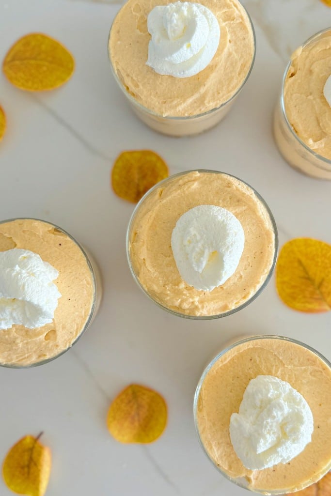 A top-down view of several glass cups filled with creamy, gluten free pumpkin mousse, each topped with a dollop of whipped cream. The glasses are surrounded by scattered yellow autumn leaves on a white surface.