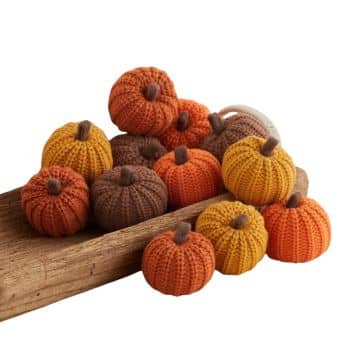 A collection of small, crocheted pumpkins in various shades of orange, yellow, and brown is displayed on a wooden surface. The pumpkins are intricately knit with stems on top, creating a cozy, autumnal feel perfect for festive fall decor.