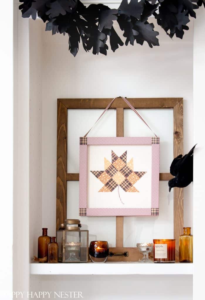 A cozy fall display featuring a framed patchwork maple leaf quilt hanging on a wooden frame, surrounded by various amber-colored glass bottles, candles, and jars. Black paper leaves and a paper raven add a Halloween touch to the festive fall decor on the shelf.