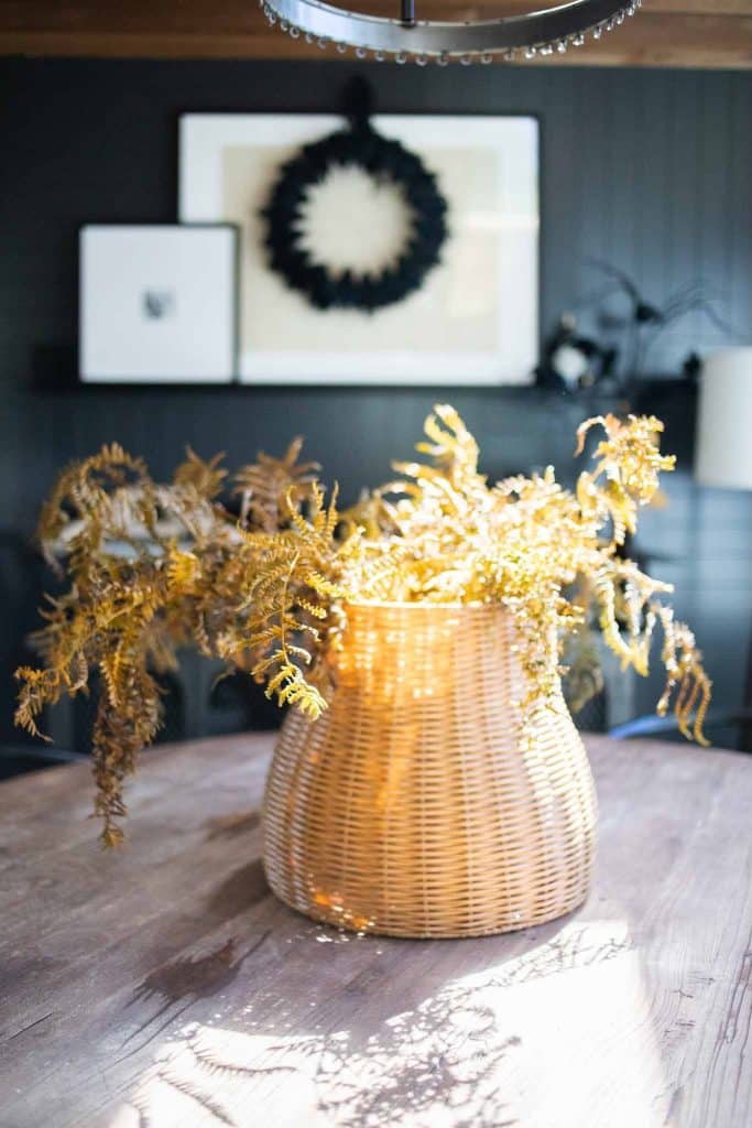 A wicker basket filled with dried golden foliage and a small pumpkin as a centerpiece sits on a wooden table in a warmly lit room. In the background, a black wreath and framed artwork are displayed on a shelf. The room has dark-toned walls and a vintage chandelier hanging above.