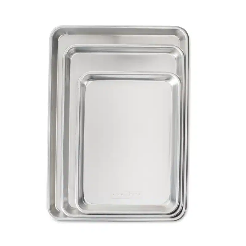 Three nested, rectangular, stainless steel baking trays of different sizes are arranged with the smallest on top and the largest at the bottom. Each tray has a slightly raised edge. They all have smooth, shiny surfaces and clean, simple designs.