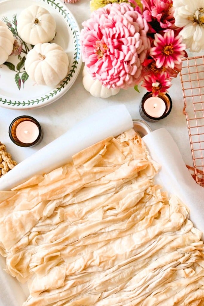 A baking tray with crinkled phyllo dough is surrounded by a floral arrangement, small white pumpkins, lit candles, and a decorative plate with more white pumpkins. The scene has a cozy, autumnal feel.