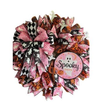 A wreath with pink and black ribbon accents featuring white ghosts and pumpkin designs. There is a round sign in the wreath that says "Spooky Season" with a ghost illustration on it. The overall design has a Halloween theme.