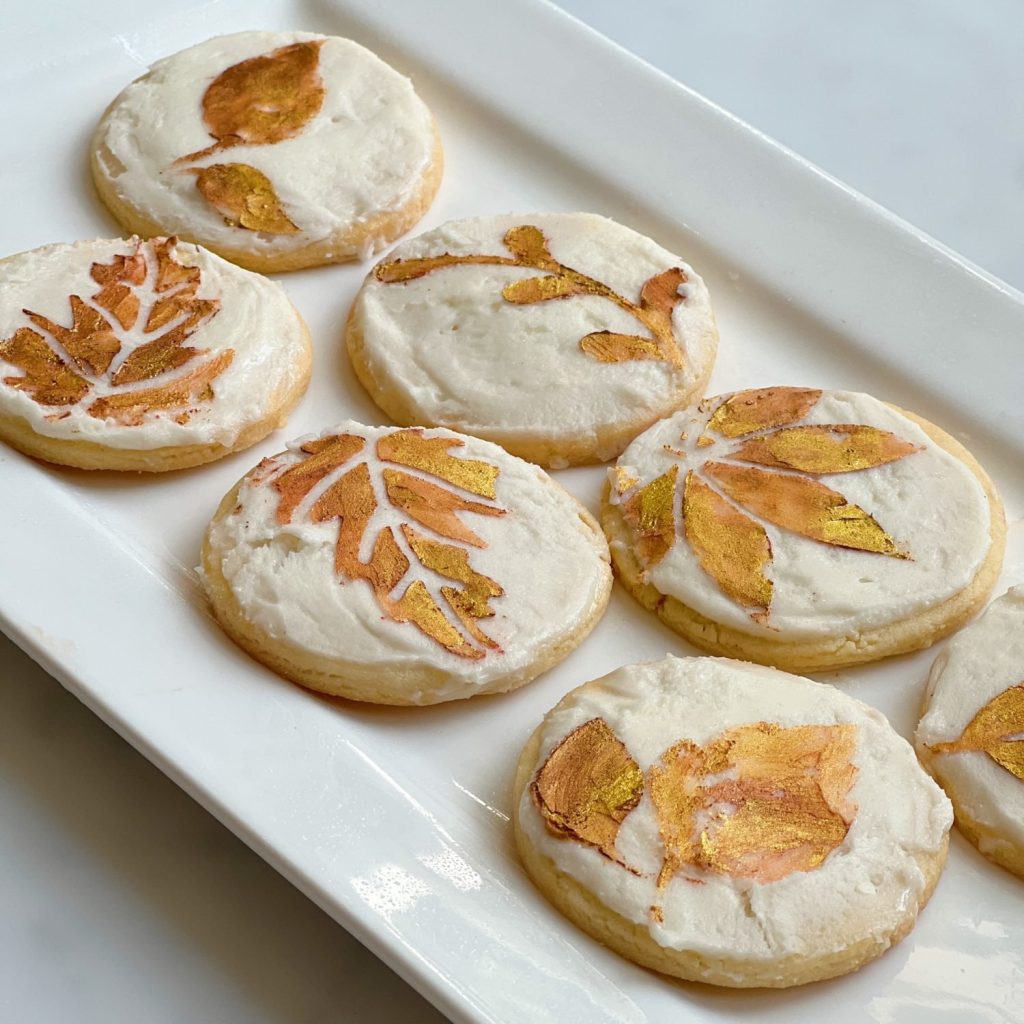 A rectangular white plate holds six round autumn cookies with white icing. Each cookie is decorated with intricate, autumn-themed leaf designs in shades of gold and bronze, showcasing different types of leaves. The background is a light, neutral surface.