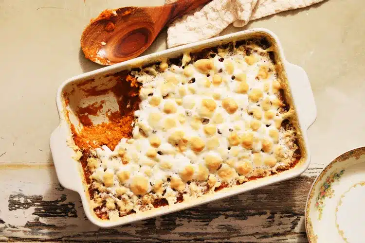 A baked sweet potato casserole topped with browned mini marshmallows in a white rectangular dish, with a serving missing from one corner. A wooden spoon with some of the filling on it rests nearby on a light-colored surface, making it perfect for your favorite autumn recipes.