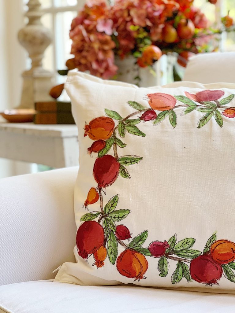 A white cushion adorned with a colorful pomegranate embroidery design is placed on a light-colored couch. In the background, a wooden table showcases fall decor ideas with a vibrant floral arrangement featuring red and orange flowers and pomegranates.