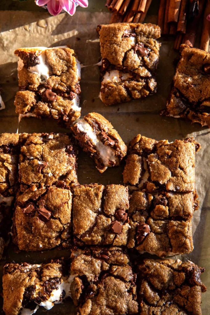 A batch of freshly baked autumn cookie bars with gooey marshmallow and melted chocolate chunks, arranged on a parchment-lined surface. A small stack of cinnamon sticks and a pink flower are visible in the corner, adding a touch of color to the warm, appetizing scene.