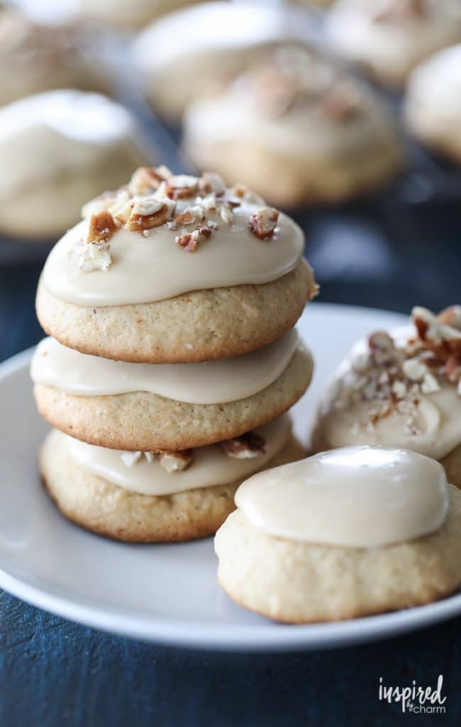 A stack of soft fall cookies with tan frosting and chopped nuts sits on a white plate, with more cookies in the background.