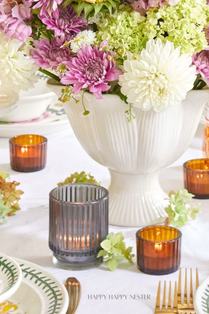 A close-up of a beautifully decorated table shows a large white vase brimming with lush flowers in shades of white, pink, and green. Surrounding the vase are several lit candles in ribbed glass holders, creating a cozy and elegant ambiance. Plates and silverware are neatly arranged nearby.