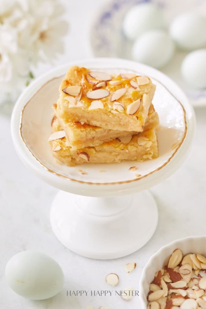 Autumn recipes: Slices of almond-topped bars arranged on a small white pedestal plate. The background includes white flowers, light blue eggs, and a small bowl filled with almond slivers.