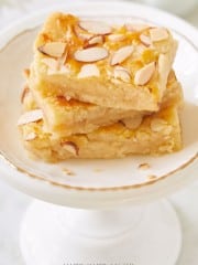 A plate holding three stacked almond-topped lemon bars. These dessert bars have a golden-yellow color and are decorated with sliced almonds on the surface. The plate is white with a gold rim and placed on a white pedestal stand.