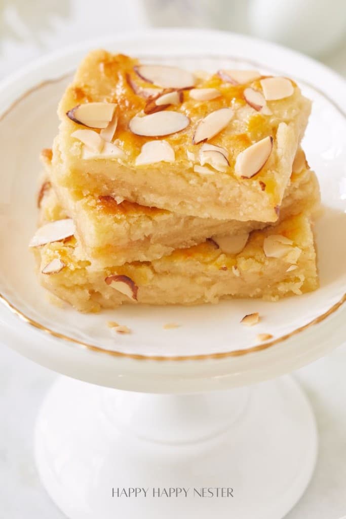 A plate holding three stacked almond-topped lemon bars. These dessert bars have a golden-yellow color and are decorated with sliced almonds on the surface. The plate is white with a gold rim and placed on a white pedestal stand.