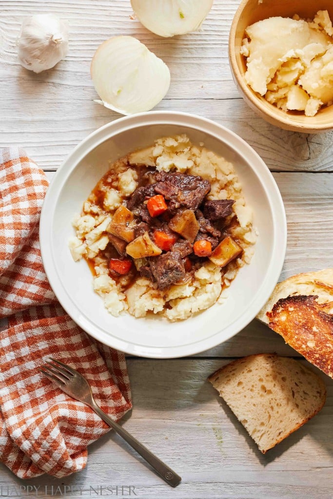 A bowl of beef stew with carrots and potatoes served over mashed potatoes graces a white wooden table. An autumn recipe favorite, it's surrounded by a striped orange and white napkin, a fork, sliced bread, chopped onions, garlic, and a bowl of grated cheese.