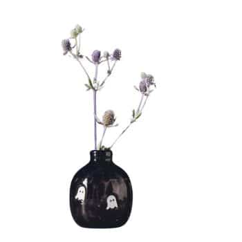 A small black vase with two ghost illustrations on its surface holds light purple thistles, arranged in a minimalist style against a white background.