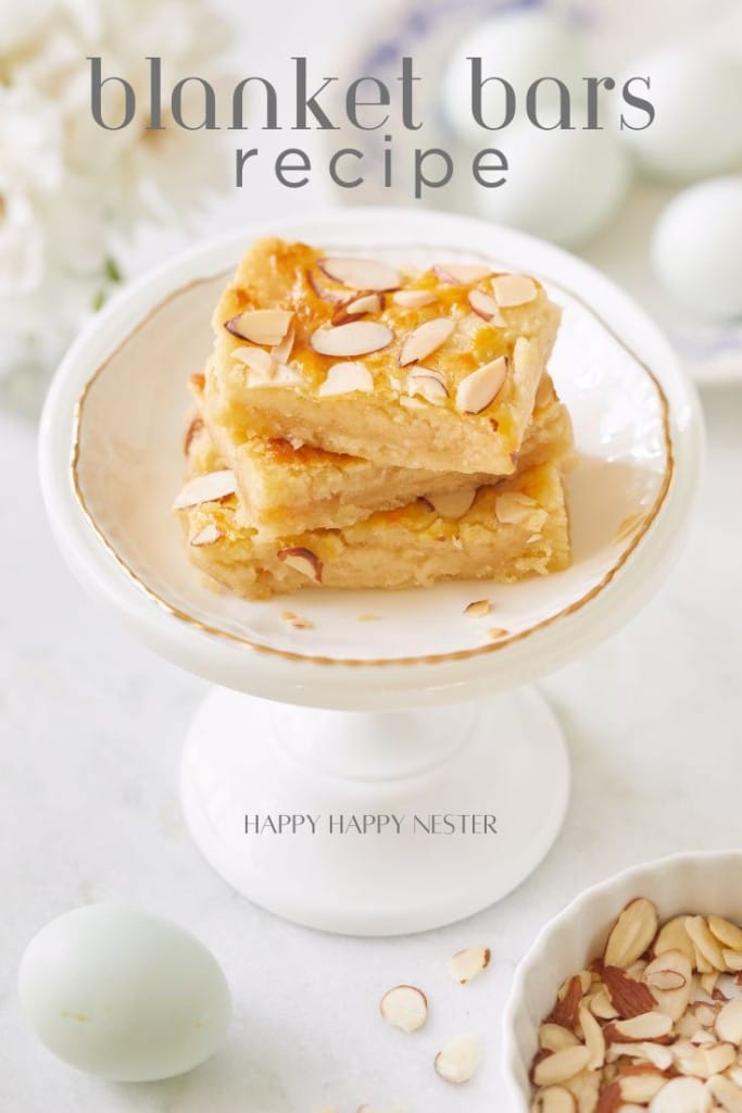 A white cake stand holds a stack of three golden brown dessert bars topped with sliced almonds. The background includes a few scattered almond slices, a light blue egg, and white flowers. Text reads "blanket bars recipe" at the top and "HAPPY HAPPY NESTER" at the bottom.