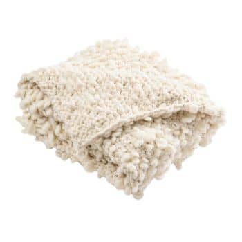 A folded, chunky knit blanket in an off-white color. The blanket has a soft and textured appearance with thick yarn loops, giving it a cozy and inviting look.