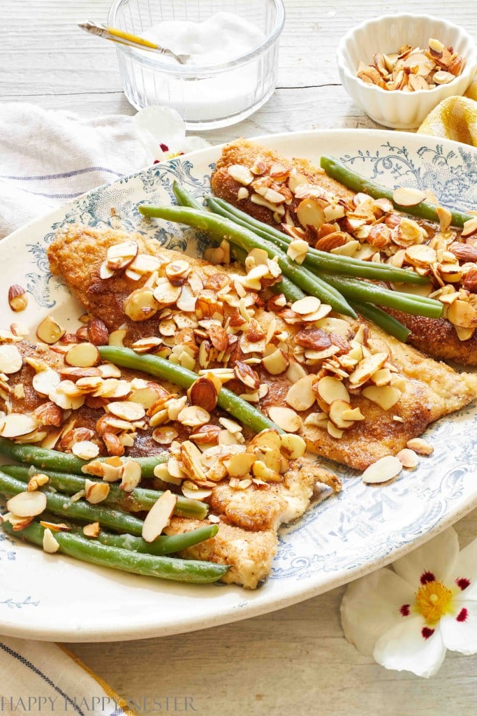 A white platter with blue floral designs holds breaded fish fillets topped with toasted almond slices—perfect for autumn recipes. Alongside the fish are fresh green beans. Also visible are a small bowl with more almond slices and a dish with a spoon, all set on a light wooden surface.