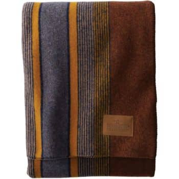 A folded woolen blanket featuring broad horizontal stripes in shades of gray, mustard yellow, and brown. A sewn-on leather patch near the bottom displays the brand's logo. The blanket has a rustic, cozy appearance.