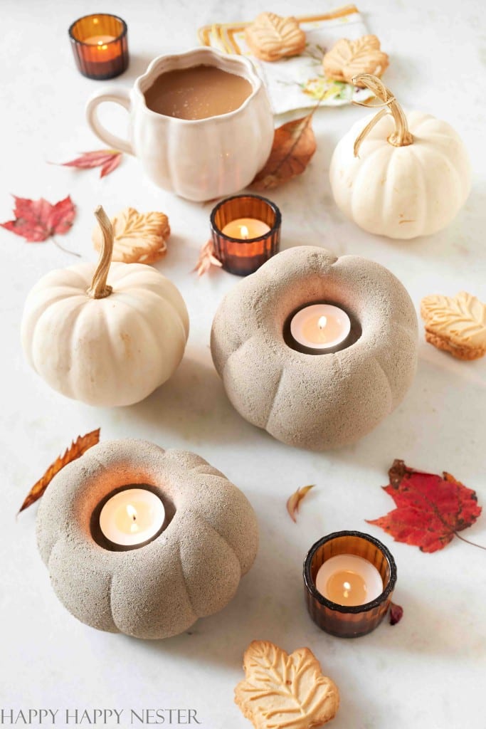 A cozy autumn scene with white pumpkins, stone-like pumpkin-shaped candle holders with lit candles, amber-colored glass tealight holders, a ceramic mug of coffee, and scattered fall leaves and leaf-shaped cookies on a white surface embodies festive fall decor.