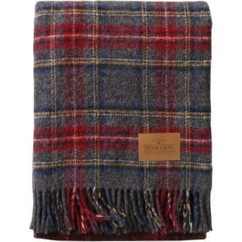 A folded, fringed wool blanket featuring a classic plaid pattern with red, gray, blue, and yellow tones. A leather patch with embossed text and logo is sewn on one side of the blanket.