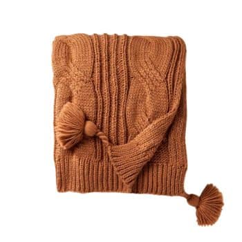 A folded, chunky knit, rust-colored sweater with cable-knit detailing and large, round tassels on the ends of a scarf-like extension.