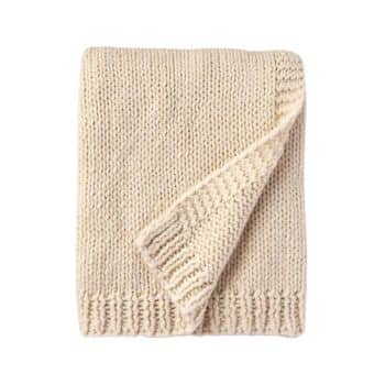 A folded, cream-colored knit blanket with visible ribbed edges, neatly arranged on a plain white background. One corner is slightly folded over, showcasing the texture and stitching detail.