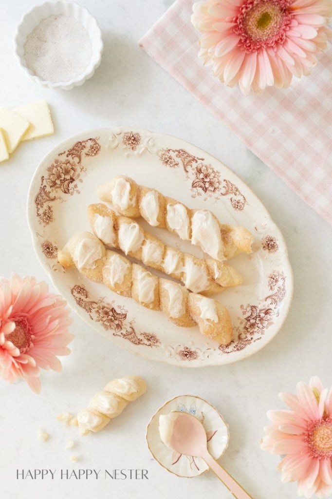 An ornate plate with three croissant twists topped with icing takes the spotlight. Surrounding the plate are pink flowers, a pink checkered cloth, a bowl with white powder, a few butter slices, and a golden spoon. The image exudes an autumn recipes vibe within its delicate, pastel-themed arrangement.