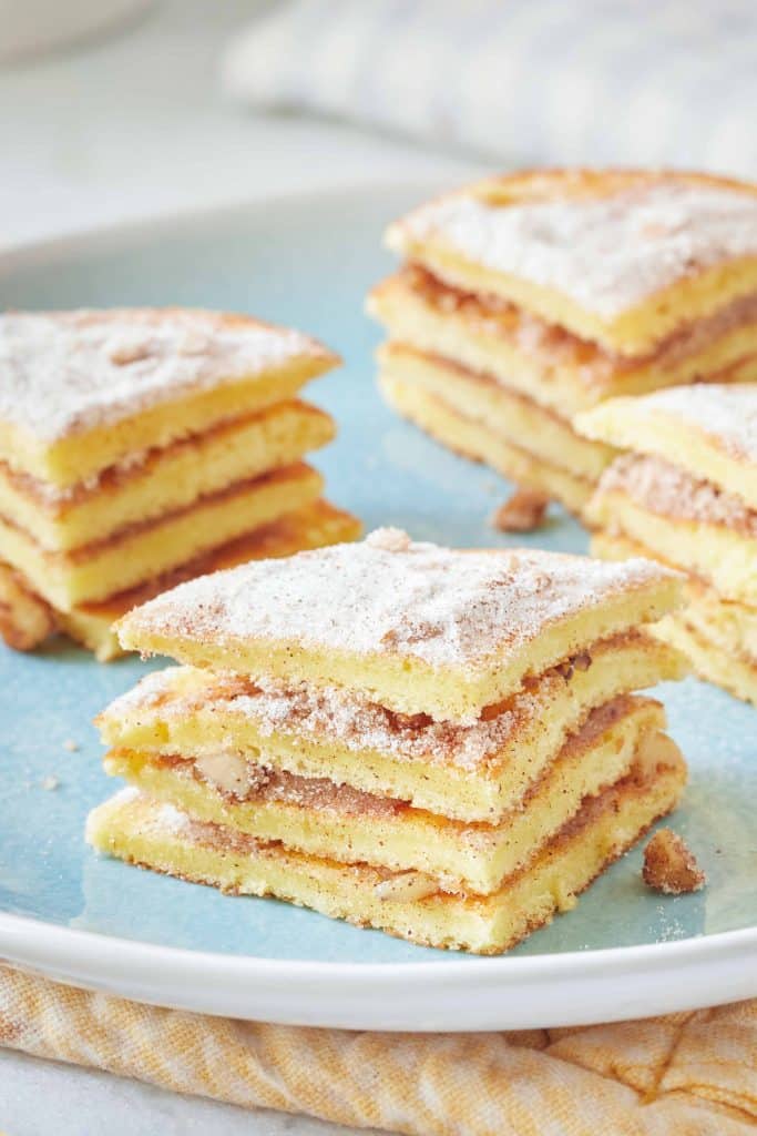 A close-up of a plate featuring several neatly stacked pastry squares. The pastries have golden layers with a light dusting of powdered sugar on top, appearing flaky and delicious—a perfect treat for autumn recipes. The plate has a soft blue hue, and a beige fabric is partially visible underneath it.