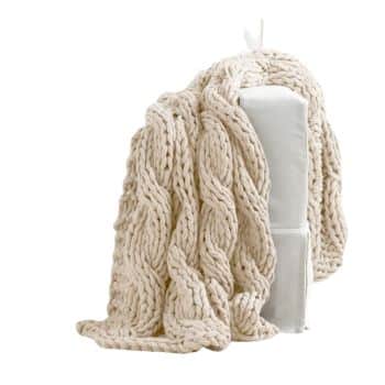 A cozy, chunky-knit blanket in a soft, neutral beige color drapes over the back of a white upholstered chair. The blanket features a large cable knit pattern, adding texture and warmth to the scene.