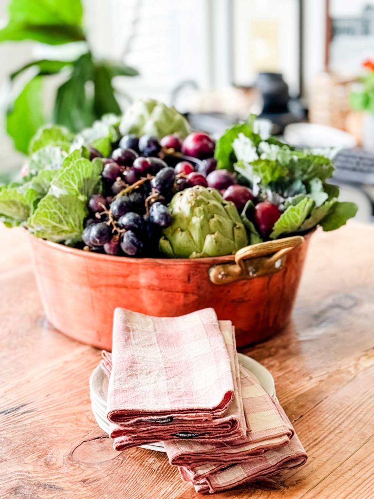 A copper bowl filled with fresh produce including grapes, artichokes, and leafy greens sits on a wooden table, making the perfect centerpiece. In front of the bowl, there are several folded pink and white checkered napkins on a small white plate. The background shows a blurred indoor setting.