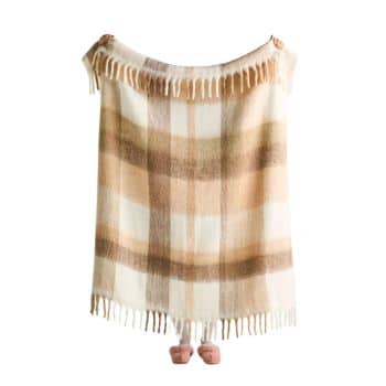 A person holding up a large, fringed, beige, brown, and cream plaid blanket. Only the person's hands and feet are visible, peeking from behind the blanket. The background is plain white.