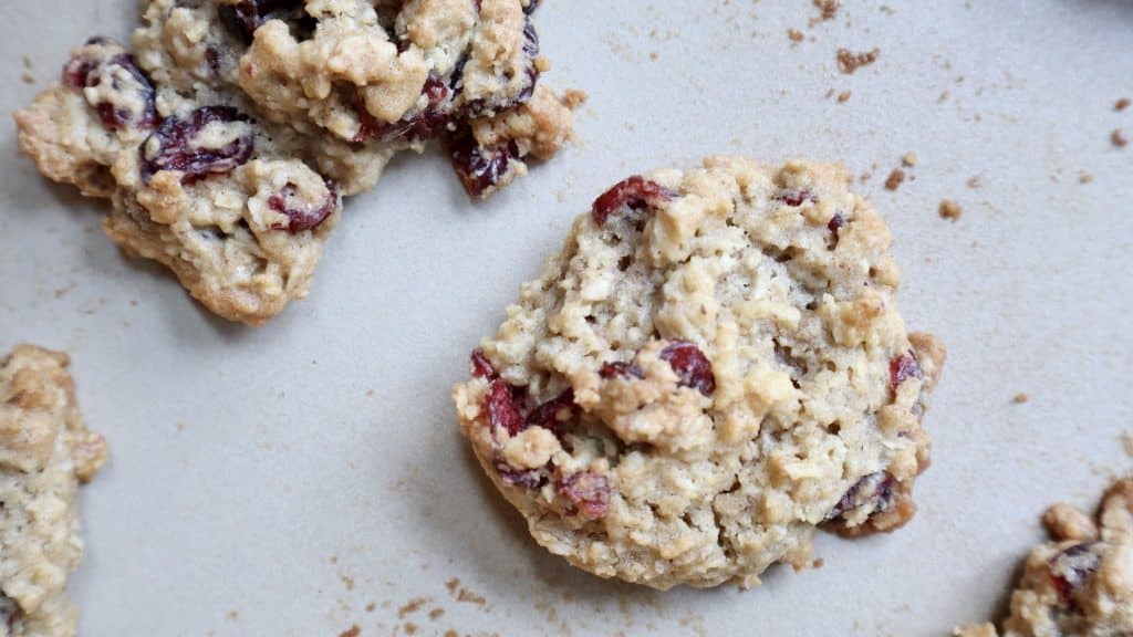 Three oatmeal fall cookies with visible cranberries rest on a light-colored surface, with a few crumbs scattered around them.