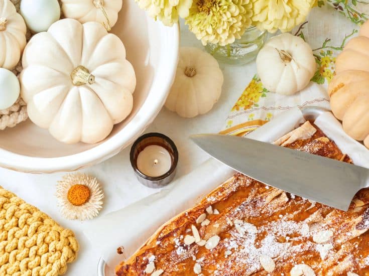 A cozy autumn scene with white pumpkins in a bowl, a lit candle, and a vase of yellow flowers. There’s a baked pastry topped with almonds and powdered sugar beside a large knife on a cutting board. A yellow knitted item and a small dried flower complete the setting.