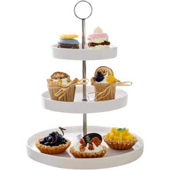 A three-tiered white dessert stand displaying an assortment of pastries inspired by autumn recipes. The top tier has a macaron and a small cake, the middle tier features two cupcakes with decorative elements, and the bottom tier has various tarts including berry, chocolate, and mango varieties.