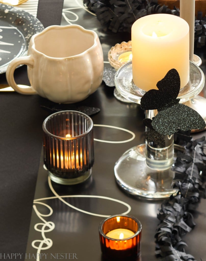 A table decorated with black, white, and amber candle holders, lit candles, a white pumpkin-shaped mug, and black paper streamers. A glittery black butterfly sits on a glass candlestick, adding a touch of elegance to the black and gold Halloween decor in this spooky-themed setup.