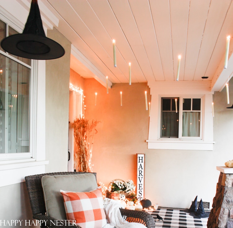 A cozy porch decorated for Halloween features festive fall decor, including a floating witch hat, suspended candles, and a "HARVEST" sign. The seating area has a wicker chair with an orange plaid cushion and a knit throw. There are also cornstalks, pumpkins, and a small stone fireplace.